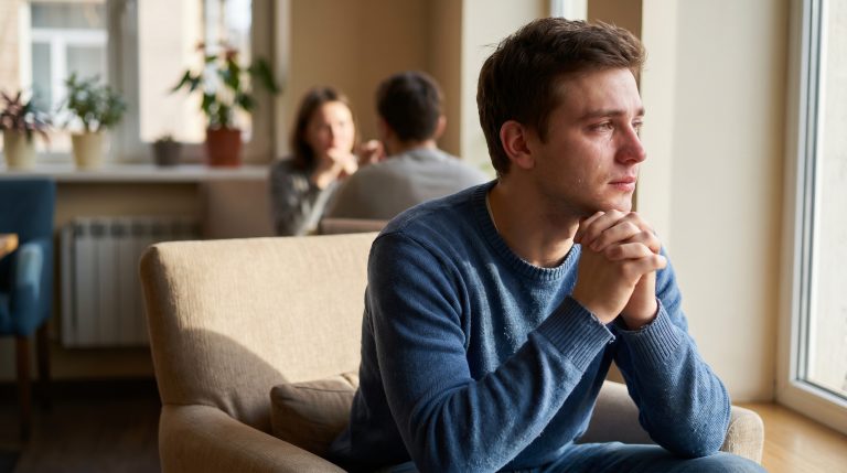 A young man sitting quietly after a difficult conversation, with people softly blurred behind him.